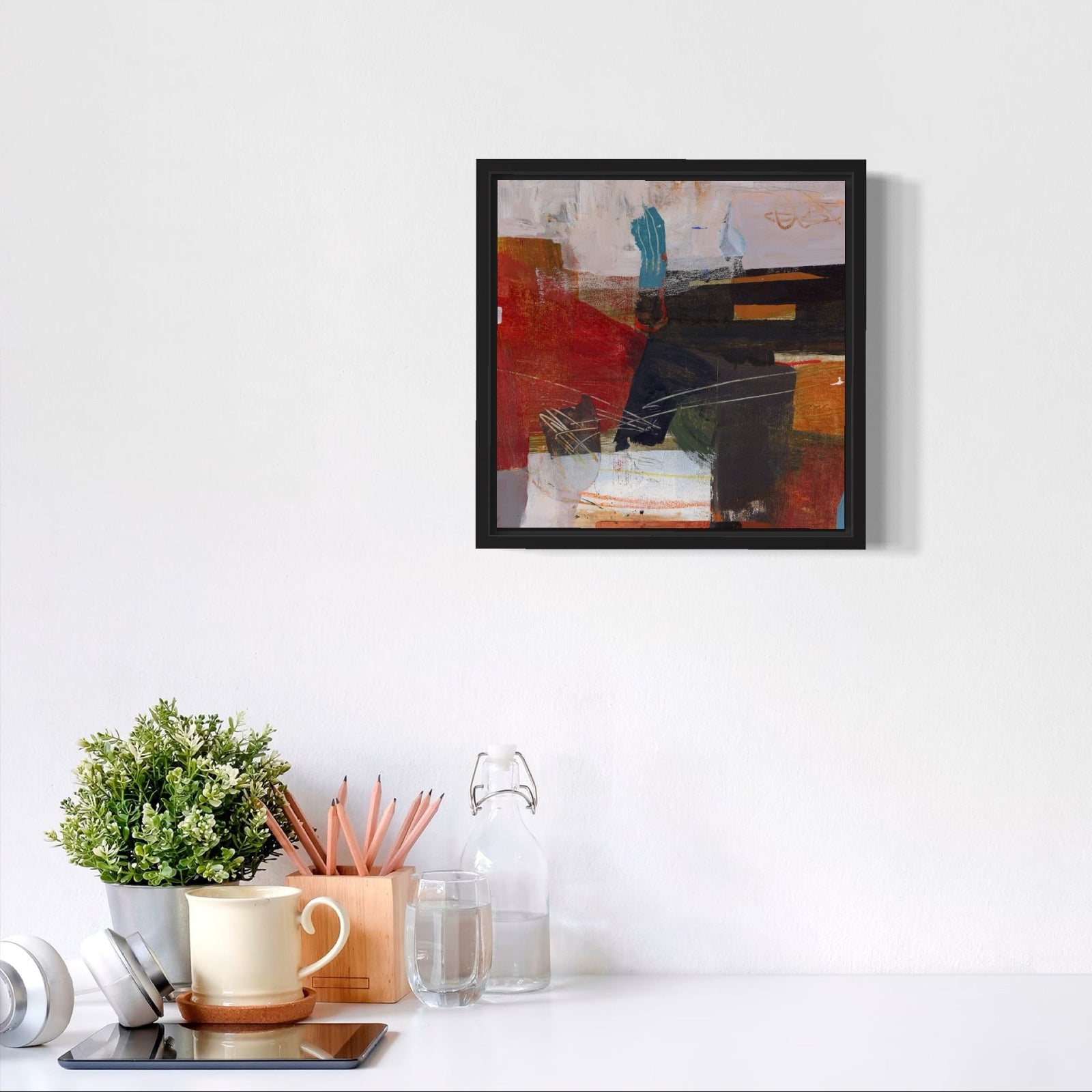 Framed abstract art piece on a white wall above a desk with office supplies.