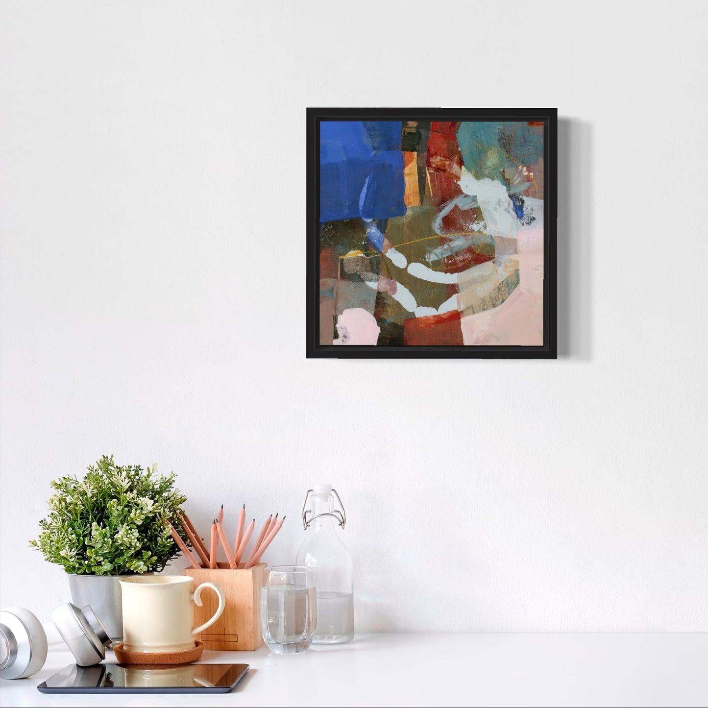 Framed abstract painting on a white wall above a desk with office supplies.