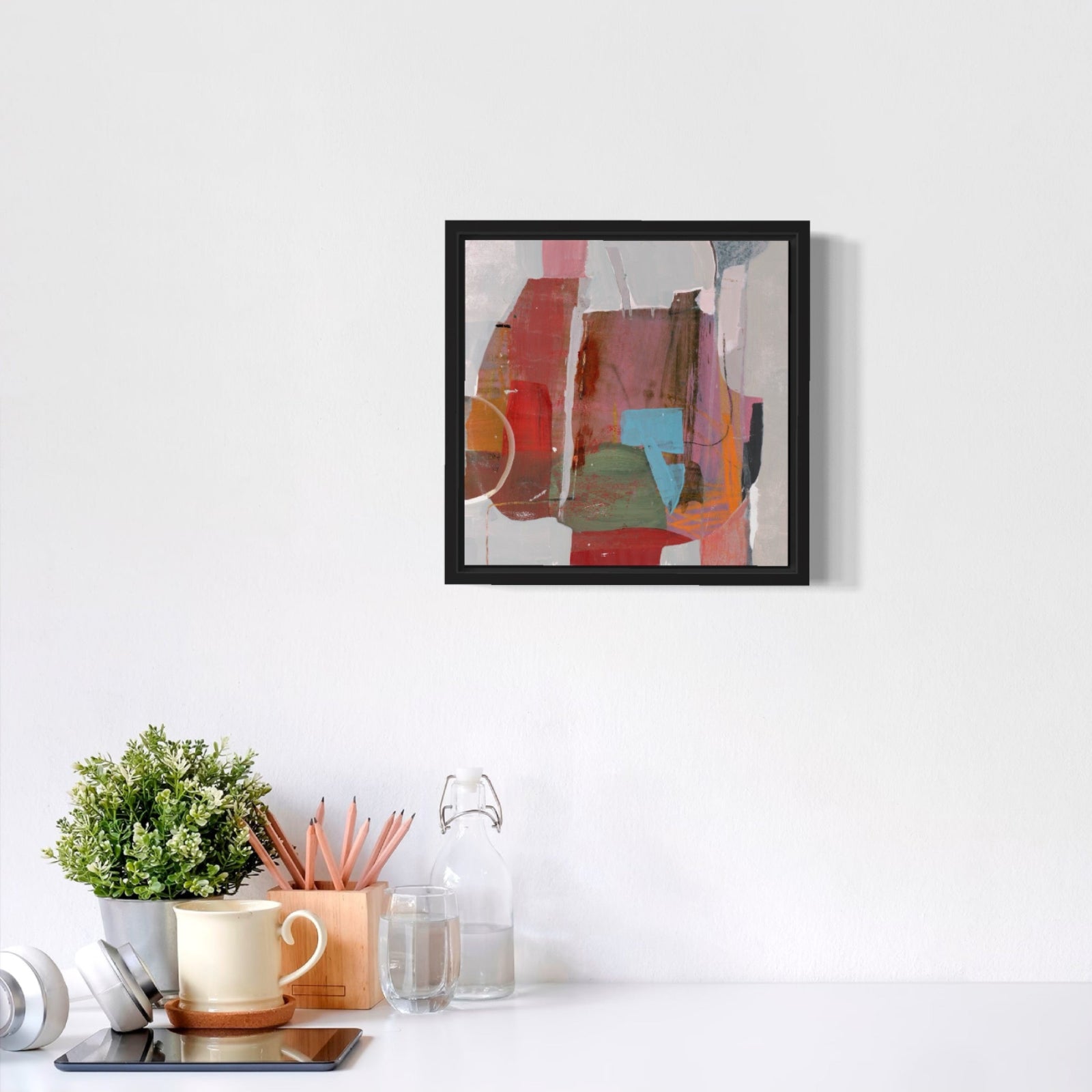 Framed abstract art piece on a white wall above a desk with office supplies.
